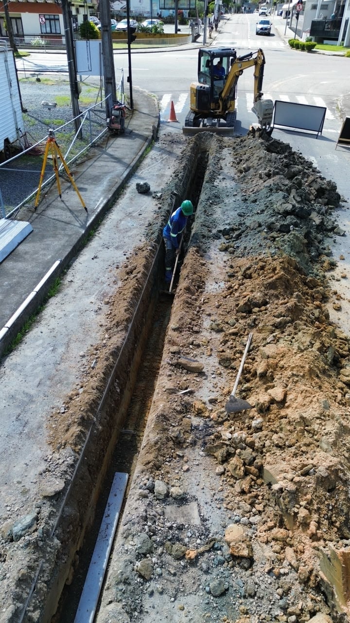 Construction site with deep trench, worker in blue, mini excavator, and traffic control equipment on urban street