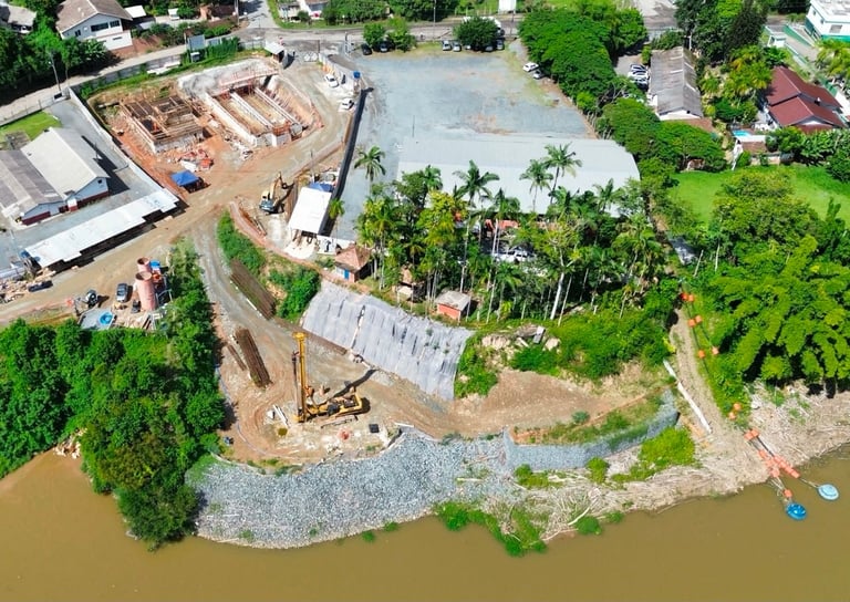 Aerial view of waterfront construction site with excavator, stockpiled materials, and partially built structures among palm trees and surrounding development